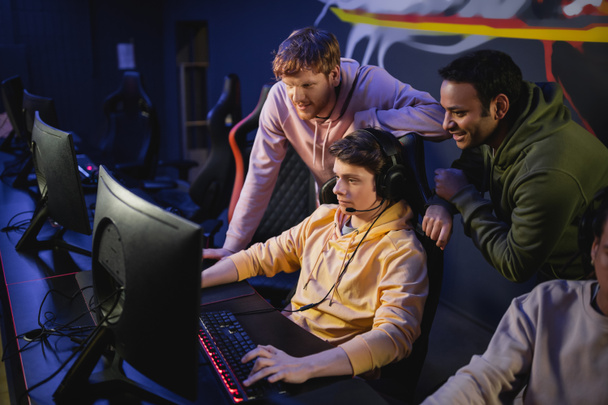 Multiethnische Freunde stehen in der Nähe von Spielern mit Kopfhörern und spielen Videospiele am Computer im Cyber-Club  - Foto, Bild
