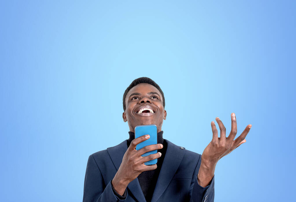 African American handsome businessman wearing formal wear is laughing holding and talking on smartphone near blue wall in background. Concept of social media, mobile application, funny joke - Photo, image