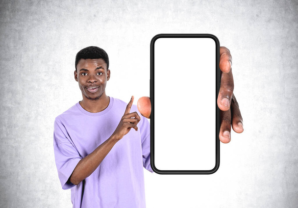 Young black man finger pointing at smartphone, large mock up copy space screen on grey concrete background. Concept of recommendation and advice - Foto, immagini