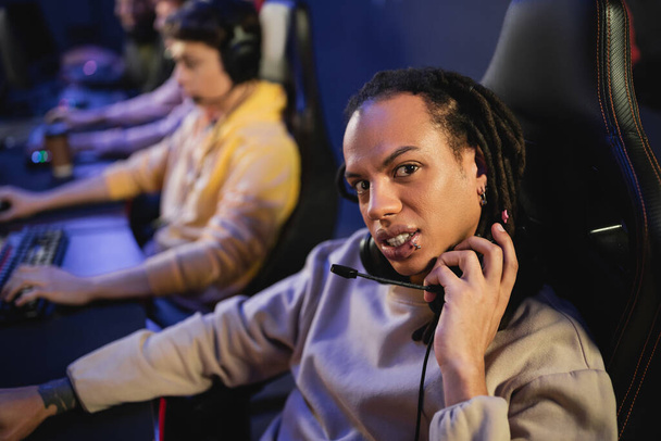 Multiraciale gamer met hoofdtelefoon en kijken naar camera in de buurt wazig team in gaming club  - Foto, afbeelding