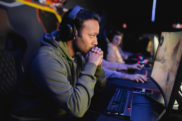 Focused indian gamer in headphones looking at computer monitor in gaming club  - 写真・画像