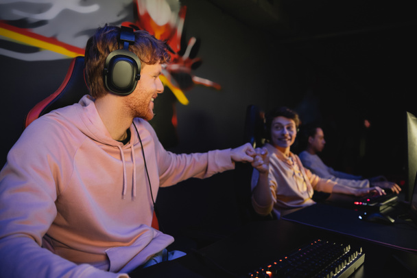 Cheerful gamer in headphones doing fist bump with blurred friend in gaming club  - Fotografie, Obrázek
