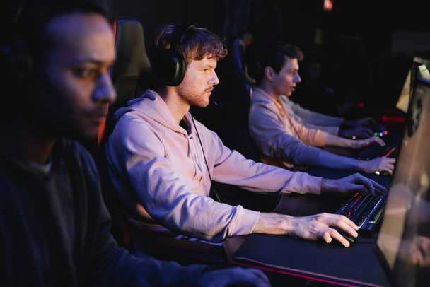 Junger Mann mit Kopfhörer spielt Videospiel auf Computer in der Nähe verschwommenes gemischtes Team in Spielclub  - Foto, Bild