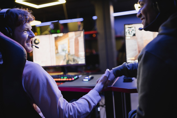 Smiling multiethnic gamers shaking hands near blurred computers in gaming club  - Photo, Image