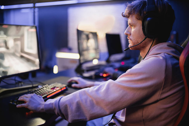 Side view of young man in headphones with microphone playing video game in cyber club  - Foto, Bild