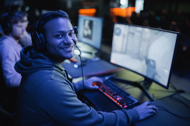 Lächelnder indischer Gamer mit Kopfhörern, der in einem Cyber-Club mit Beleuchtung in die Kamera neben dem Computer blickt  - Foto, Bild
