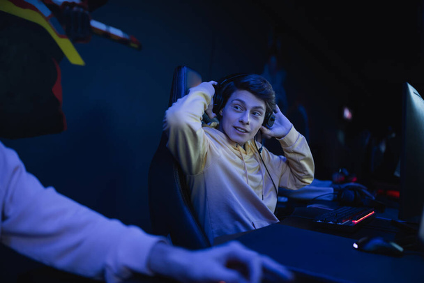 Młody gracz w słuchawkach rozmawia z niewyraźnym przyjacielem w klubie cybernetycznym z oświetleniem  - Zdjęcie, obraz