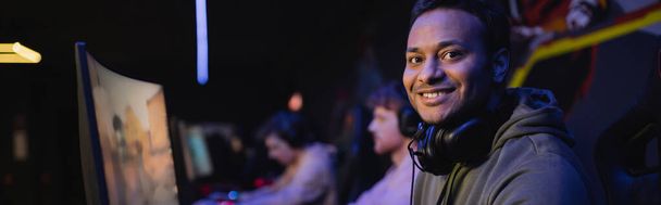 Positive indian gamer with headphones looking at camera in cyber club, banner  - Fotoğraf, Görsel