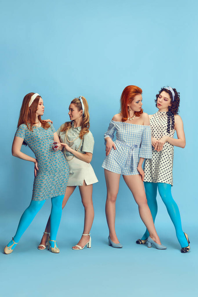 Friends meeting. Full-length portrait of attractive young women posing in stylish clothes against blue studio background. Concept of retro style, fashion, beauty, elegance, 60s, youth. Pin-up style - 写真・画像