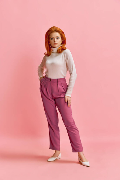 Full-length portrait of beautiful redhead woman in stylish clothes, hairdo and makeup posing against pink studio background. Concept of retro style, fashion, beauty, elegance, 60s, youth. Pin-up style - Foto, Imagen