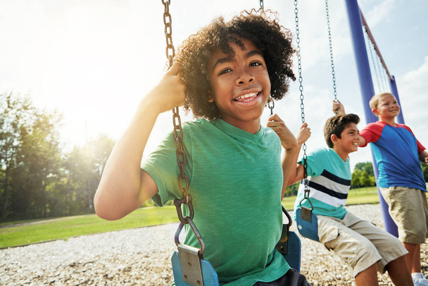 Play is vital for a kids healthy development. Portrait of a young boy playing on a swing at the park with his friends - Photo, Image