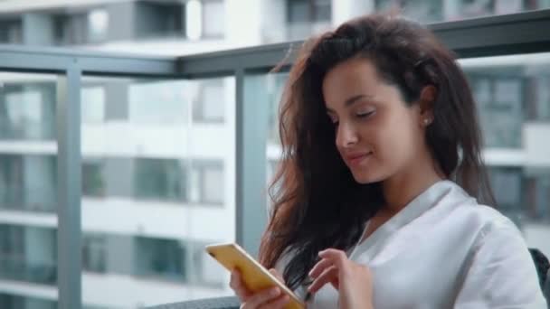Onnellinen kaunis nainen lepää parvekkeella kotona pitäen matkapuhelin, kirjoittamalla viestin. Nainen silkkiaamutakissa. Ajatus aamusta nauttimisesta. Elämäntapa. Reaaliaikainen - Materiaali, video