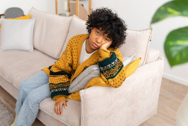 Αφρο-αμερικάνα λυπημένη σκεπτική κοπέλα που κάθεται στον καναπέ στο σπίτι. Νεαρή Αφρικανή που αναλογίζεται φαίνεται κουρασμένη μετά από μια κουραστική μέρα. Το κορίτσι αισθάνεται κατάθλιψη προσβεβλημένη μοναχική διαταραχή - Φωτογραφία, εικόνα