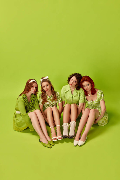Portrait of pretty, stylish young women, friends posing against green studio background. Attractive beauty. Concept of retro style, fashion, beauty, elegance, 60s, youth. Pin-up style. Top view - 写真・画像