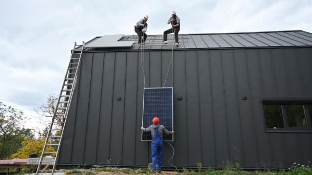 Muži instalují solární panely na střechu domu. Elektrikáři v helmách zvedají fotovoltaický solární modul pomocí lan venku. Koncepce alternativní a obnovitelné energie. - Záběry, video