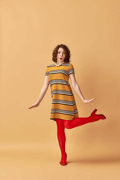 Full-length portrait of emotive brunette young woman with curly hair, in stylish clothes posing against orange studio background. Concept of retro style, fashion, beauty, elegance, 60s. Pin-up style - 写真・画像