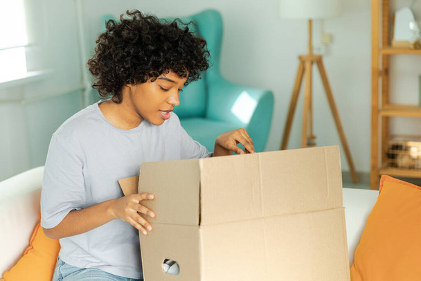 African girl unpacking delivery looking in box. Happy woman opening carton box. Female getting parcel looking at delivered goods items. Satisfied client positive feedback quick express deliver - Foto, Bild