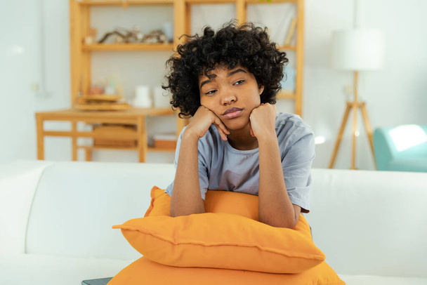 Αφρο-αμερικάνα λυπημένη σκεπτική κοπέλα που κάθεται στον καναπέ στο σπίτι. Νεαρή Αφρικανή που αναλογίζεται φαίνεται κουρασμένη μετά από μια κουραστική μέρα. Το κορίτσι αισθάνεται κατάθλιψη προσβεβλημένη μοναχική διαταραχή - Φωτογραφία, εικόνα
