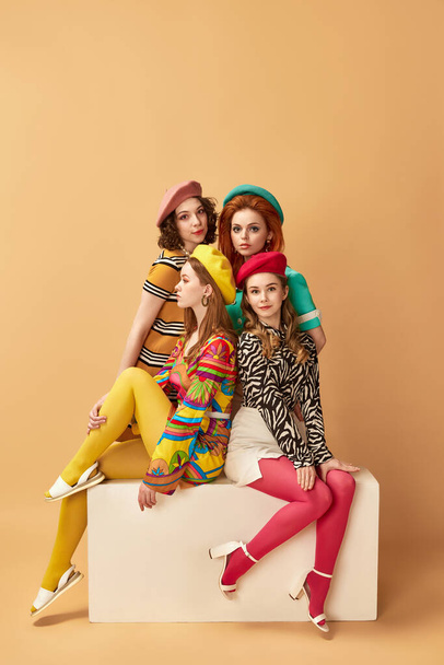 Ritratto di giovani donne attraenti con trucco, acconciatura elegante e vestiti colorati che posano sullo sfondo dello studio arancione. Stile retrò, moda, bellezza, eleganza, anni '60, concetto di gioventù. Stile pin-up - Foto, immagini