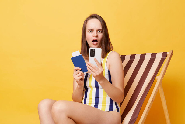 Shocked amazed astonished woman wearing striped swimming suit sitting on deck chair isolated over yellow background posing with passport, using mobile phone. - Photo, Image
