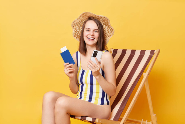 Cheerful caucasian adult woman wearing striped swimming suit sitting on deck chair isolated over yellow background using cell phone holding passport booking tickets abroad. - Фото, зображення