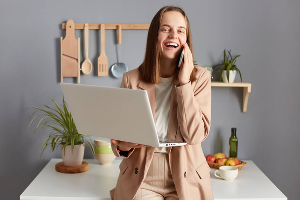 Extremely happy woman with brown hair wearing formal suit posing in kitchen, working from hone on notebook , having break, talking on smart phone. - Photo, Image