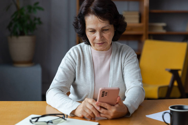 Ευρωπαϊκή μεσήλικη ηλικιωμένη γυναίκα που κατέχει χρησιμοποιώντας το smartphone οθόνη αφής πληκτρολογώντας κύλιση σελίδα. Κομψή ηλικιωμένη ώριμη 60άρα κυρία με κινητό τηλέφωνο που χρησιμοποιεί διαδικτυακές εφαρμογές κοινωνικής δικτύωσης στο σπίτι. Αγορές online - Φωτογραφία, εικόνα