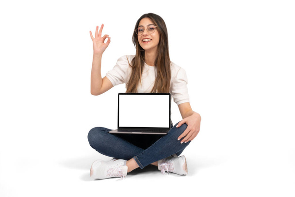 Laptop mock up, young girl sitting on the ground and holding laptop mock up. Recommending web site,  gesturing ok sign with her hand. New application, advertising, modern technology concept idea.  - Foto, Bild