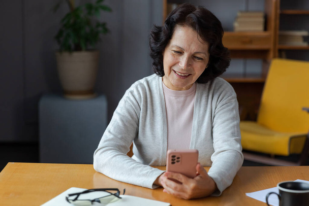 Ευρωπαϊκή μεσήλικη ηλικιωμένη γυναίκα που κατέχει χρησιμοποιώντας το smartphone οθόνη αφής πληκτρολογώντας κύλιση σελίδα. Κομψή ηλικιωμένη ώριμη 60άρα κυρία με κινητό τηλέφωνο που χρησιμοποιεί διαδικτυακές εφαρμογές κοινωνικής δικτύωσης στο σπίτι. Αγορές online - Φωτογραφία, εικόνα