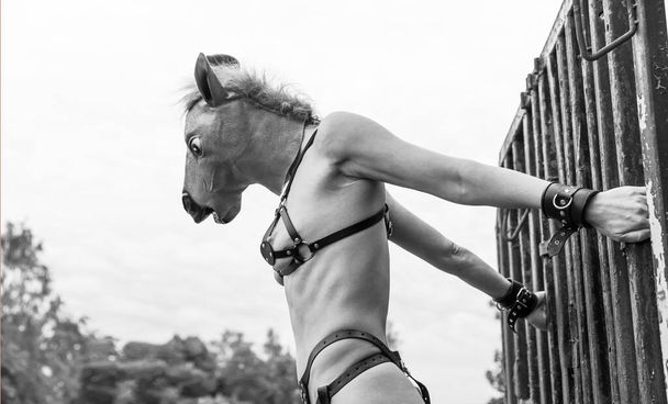 Jovem mulher nua em cintos de couro e vestindo uma máscara de cavalo posando perto de um velho trailer enferrujado. Mulher nua posando em uma lixeira de máquinas agrícolas velhas - Foto, Imagem
