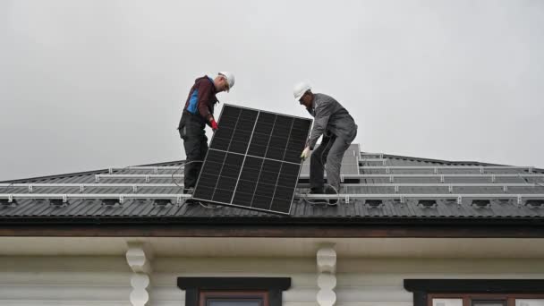 Muži instalují solární panely na střechu domu. Elektrikáři v helmách zvedají fotovoltaický solární modul pomocí lan venku. Koncepce alternativní a obnovitelné energie. - Záběry, video