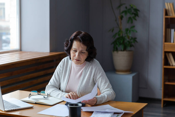 Middle aged senior woman sit with laptop and paper document. Pensive older mature lady reading paper bill pay online at home managing bank finances calculating taxes planning loan debt pension payment - 写真・画像