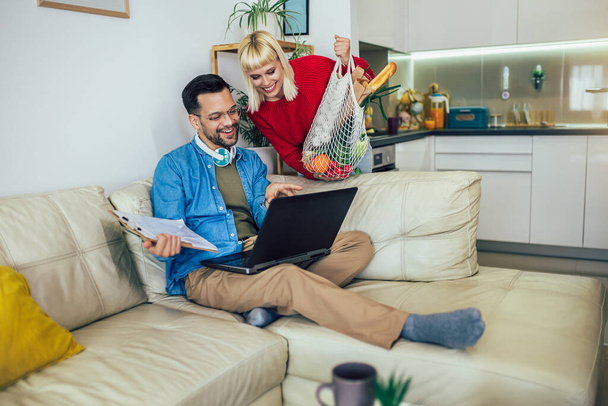 Young man is working on his laptop while wearing headphones and sitting on a comfortable couch. His wife has just arrived home from grocery shopping, holding a bag of fresh produce. - Foto, imagen