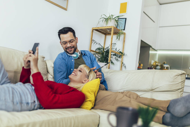 Hermosa pareja abrazándose relajante en el sofá y utilizando la tableta digital y el teléfono. - Foto, imagen