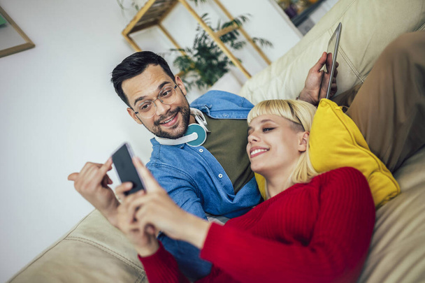 Красивая пара обнимает расслабляясь на диване и с помощью цифрового планшета и телефона. - Фото, изображение
