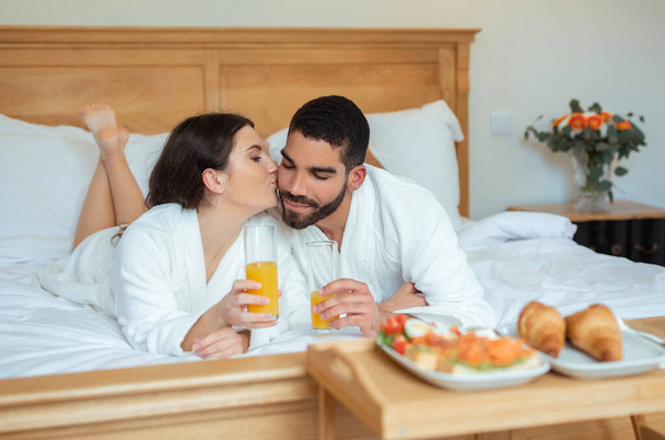 Romance de luna de miel. Esposa cariñosa besando al marido en la mejilla bebiendo jugo y disfrutando de un desayuno romántico en la cama acostado en la moderna suite del hotel interior, con albornoces. Enfoque selectivo - Foto, imagen