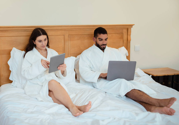 Ανεξάρτητοι Ζευγάρι Χρησιμοποιώντας Laptop Και Ψηφιακούς Υπολογιστές Tablet Που Δουλεύουν Online Και Περιήγηση Διαδικτύου Κάθονται Στο Κρεβάτι Στο Σπίτι, Φορώντας Λευκά Μπουρνούζια. Τεχνολογία και Gadgets Concept - Φωτογραφία, εικόνα