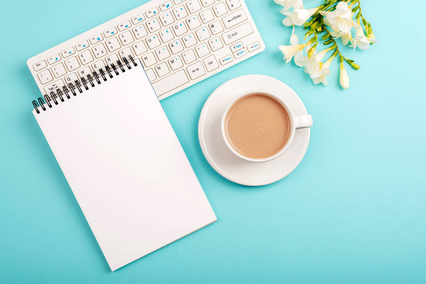 Computer keyboard, blank notepad and coffee cup on blue table with freesia flowers. Top view, flat lay, mockup. - Photo, Image