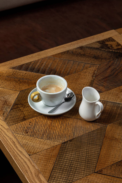 小さじ1杯のソーサーの上に軽いセラミックカップの牛乳とコーヒー。近くには軽いセラミックミルクジャグがあります。木のテーブルの上に料理がある. - 写真・画像