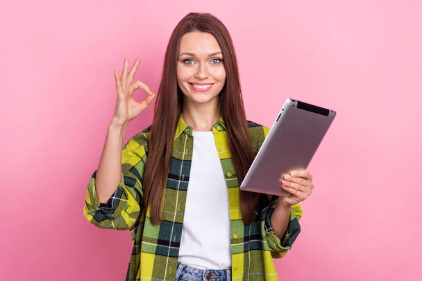 Portrait of satisfied friendly girl with straight hairdo dressed plaid shirt hold tablet showing okey isolated on pink color background. - Foto, Bild