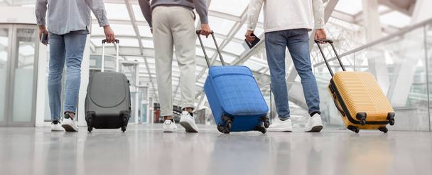 Путешествие. Обрезанный снимок трех мужчин, идущих с чемоданами обратно в камеру в терминале аэропорта внутри. Неузнаваемые друзья, парни, отправляющиеся в отпуск с багажом. Панорама, вид сзади - Фото, изображение