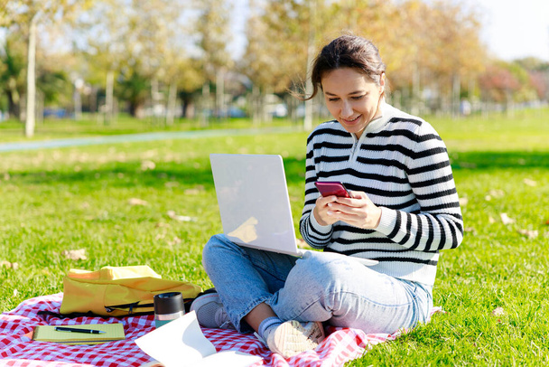 Young woman sitting in park with laptop and using smart phone - Foto, imagen