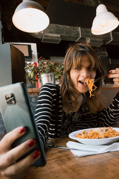 vertical image of transgender young latin woman of argentinian ethnicity eating spaghetti inside a restaurant and taking a selfie - Photo, Image