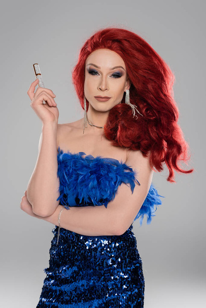 Drag queen in dress with feathers holding mascara isolated on grey   - Photo, image