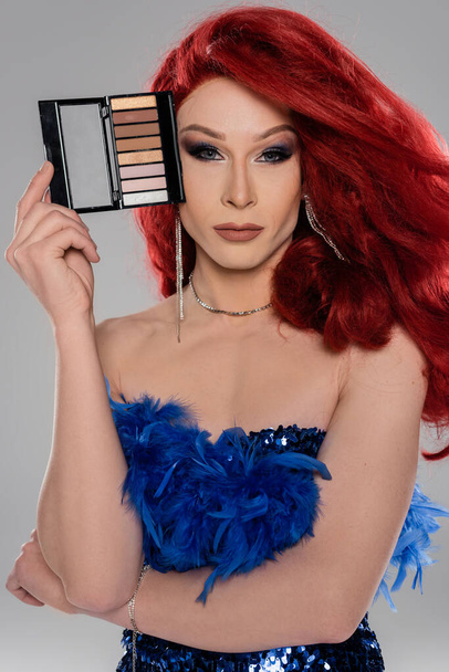 Πορτρέτο της drag queen σε κόκκινο περούκα κρατώντας σκιές ματιών και κοιτάζοντας κάμερα που απομονώνονται σε γκρι   - Φωτογραφία, εικόνα