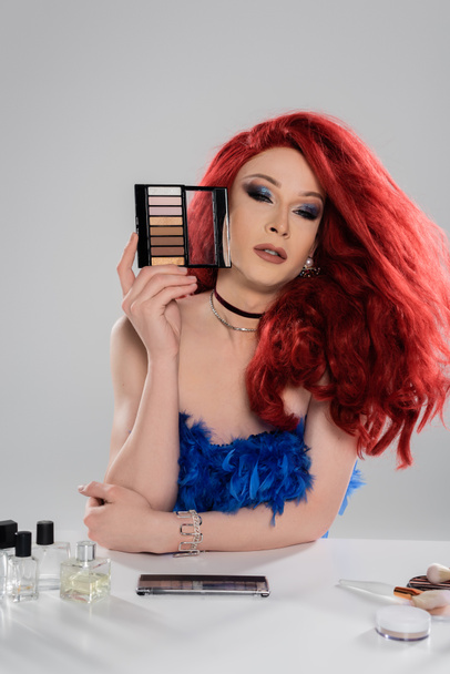 Drag queen en peluca roja sosteniendo sombras de ojos paleta cerca de perfumes y pinceles de maquillaje aislados en gris   - Foto, Imagen