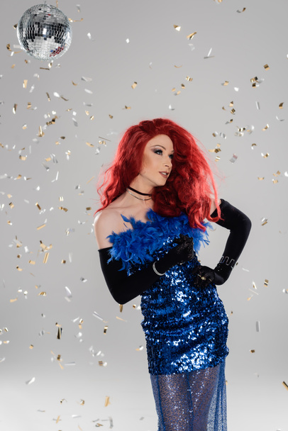 Glamorous drag queen in dress and gloves standing under confetti and disco ball on grey background  - Foto, immagini
