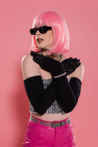 Portrait of trendy drag queen in sunglasses and gloves touching shoulders on pink background  - Foto, Bild