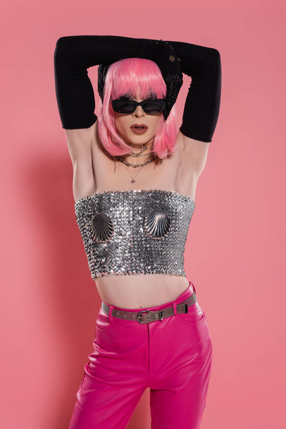 Fashionable drag queen in sunglasses and shiny top touching hair on pink background  - Photo, Image