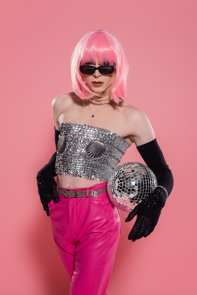Fashionable drag queen in sunglasses and gloves holding disco ball on pink background  - Foto, imagen